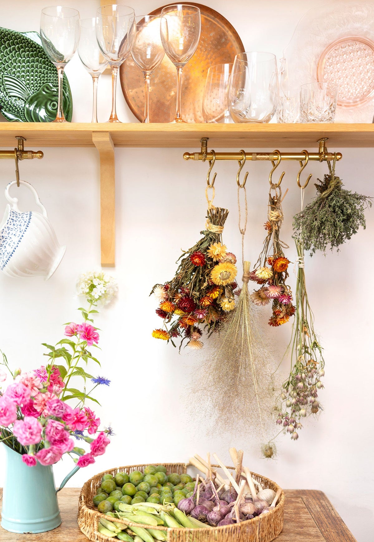 A styled kitchen or shelf display with wooden shelves holding glassware and dried flowers hanging from brass hooks. A light blue pitcher with pink flowers sits on a wooden table next to a woven basket containing green olives and purple garlic.