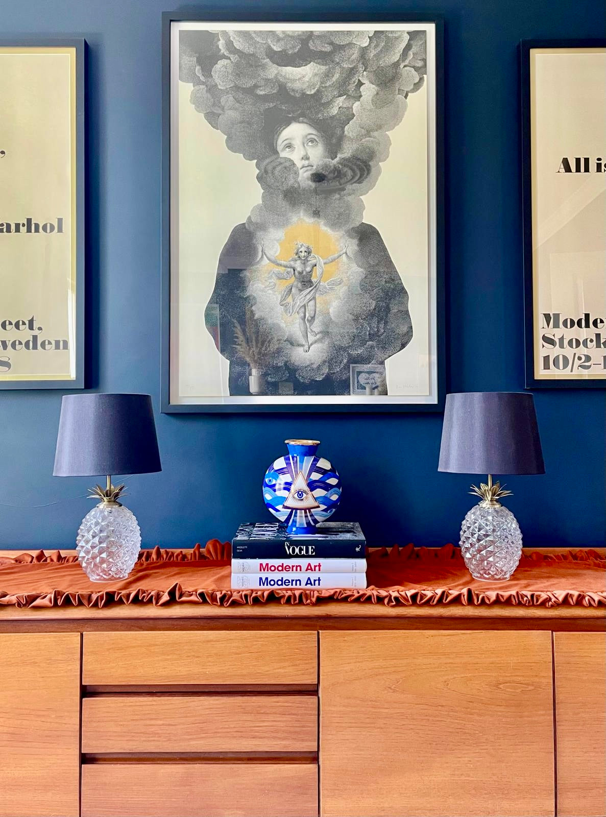 A framed art print featuring a surreal portrait of a woman whose face is obscured by smoke, displayed on a dark blue wall above a wooden console table with two pineapple-shaped table lamps and decorative books.