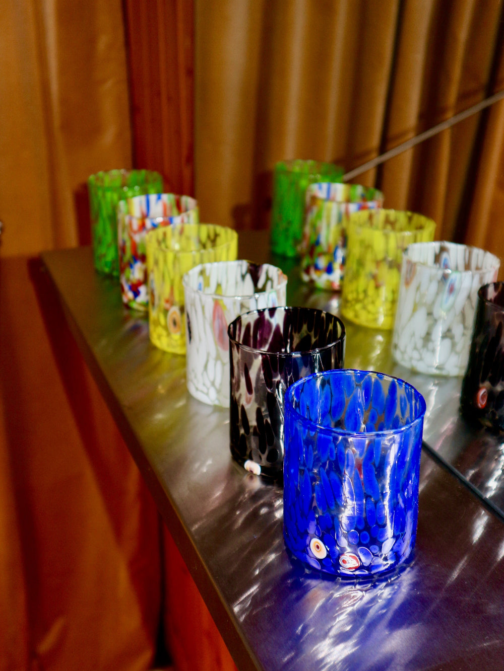 Collection of colourful Murano glass tumblers on a reflective surface with a wooden background
