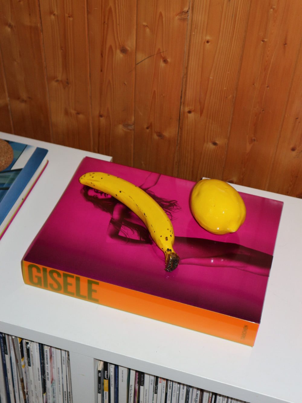 Book titled 'Gisele' with a ceramic banana and ceramic lemon on a pink surface against a wooden wall.