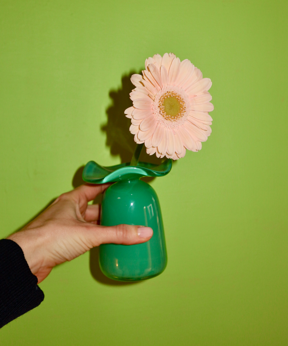 Hand holding a small green vase with a pink flower against a green background