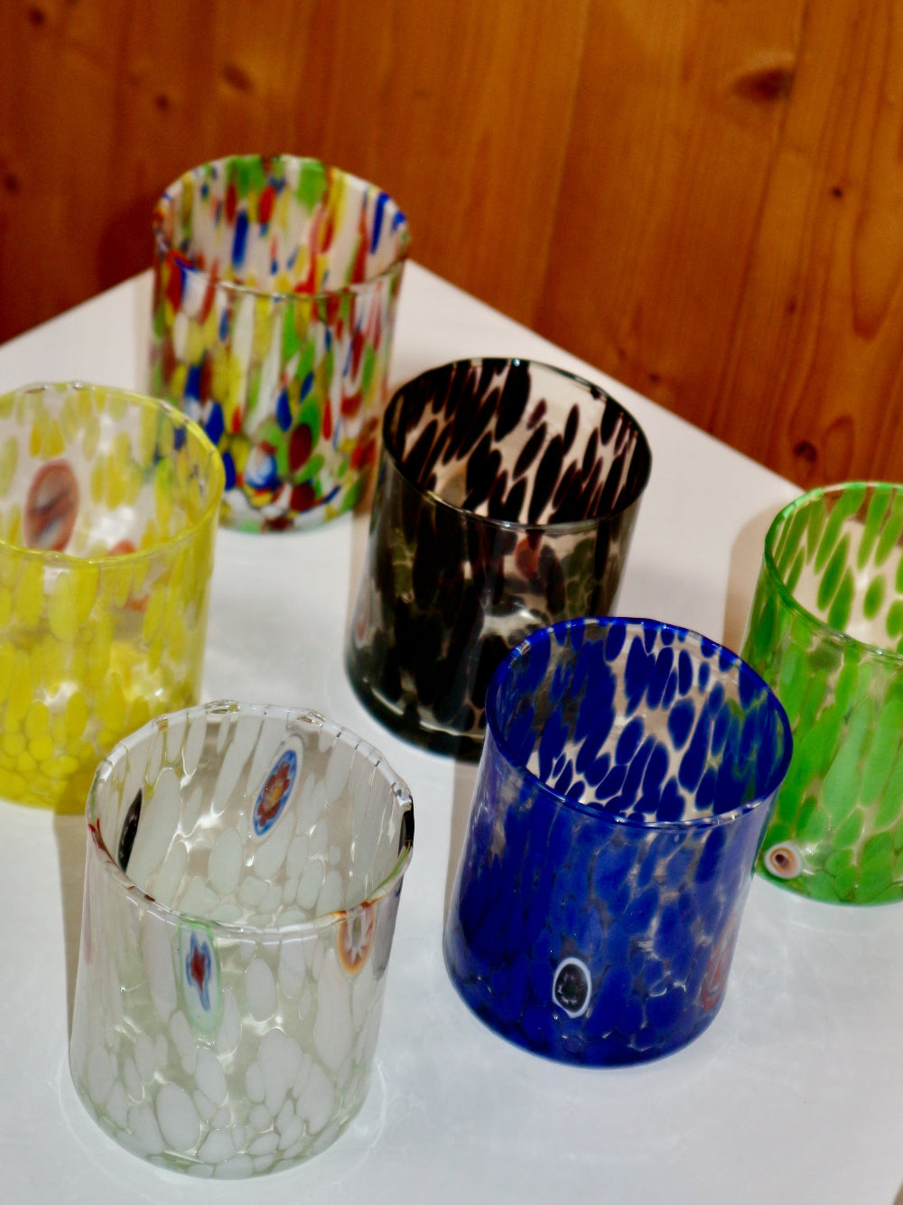 Collection of colourful murano glass tumblers on a white surface with a wooden background