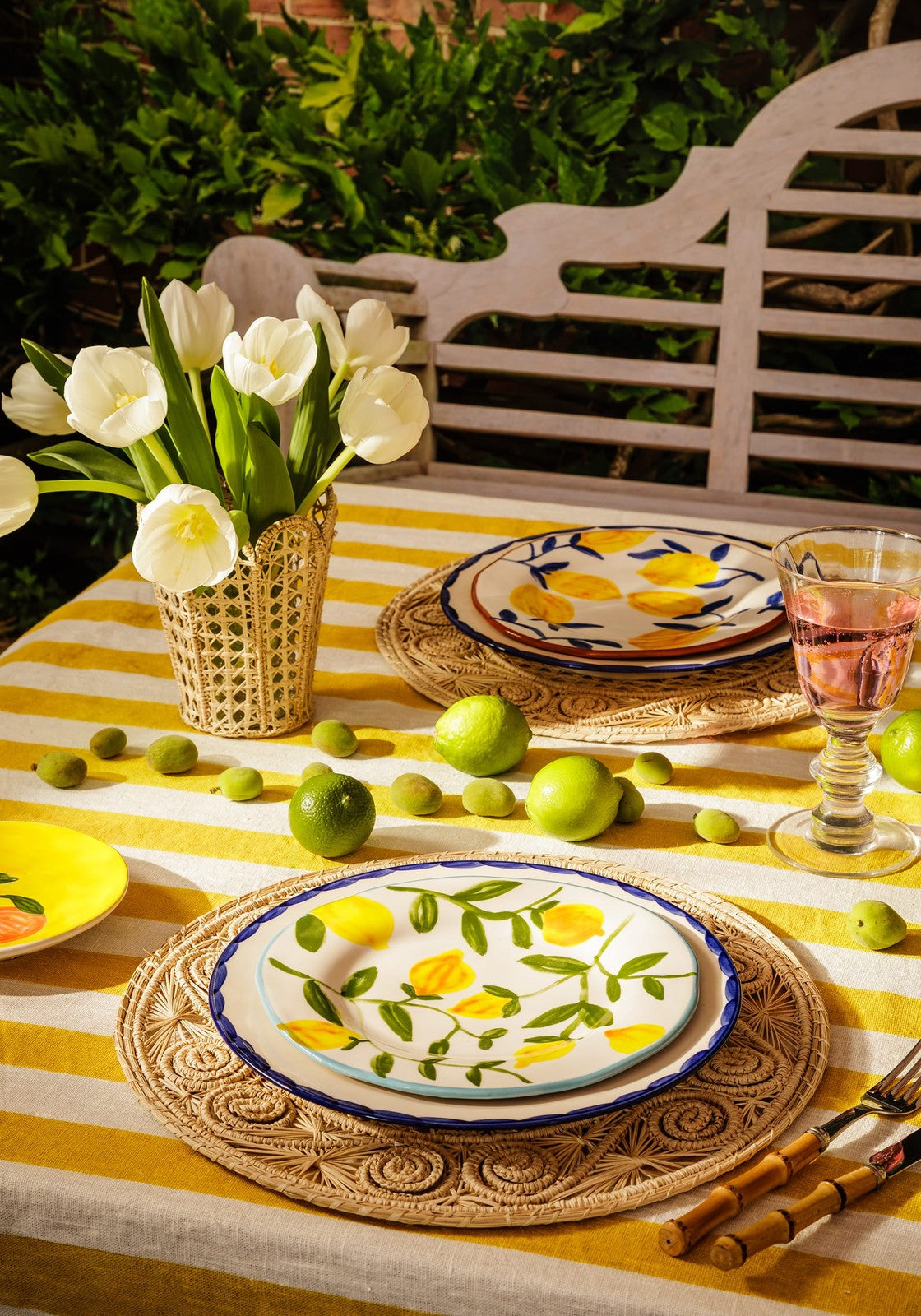 Outdoor table setting with floral plates, lemons, and a bench in the background.