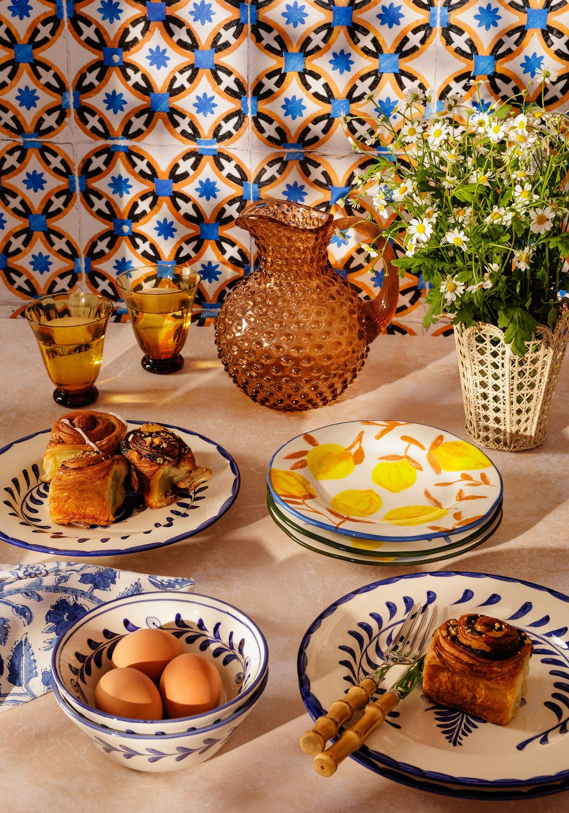 Table setting with lemon painted ceramic plates, bowls, and a vase against a patterned wall.