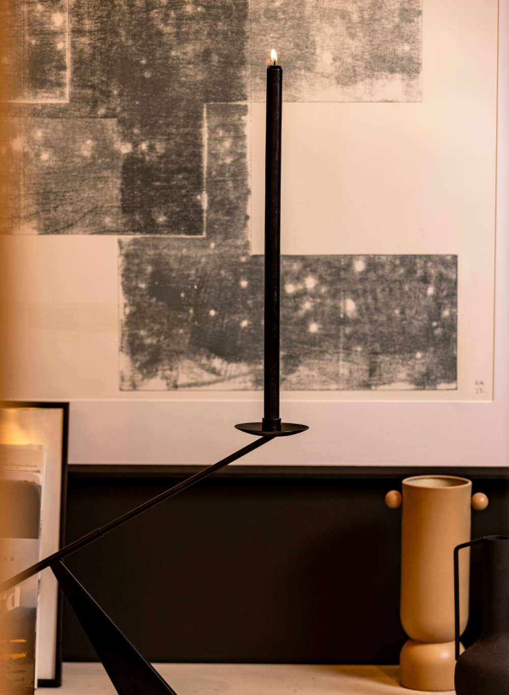 Black candlestick with a candle on a reflective surface in a room with abstract art on the wall.