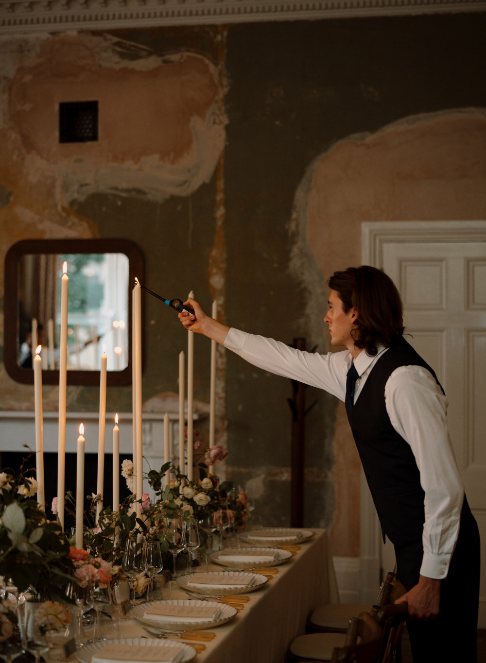 Man in formal attire lighting candles at a decorated table in an elegant room.