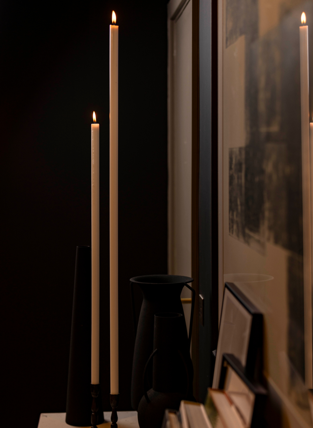 Tall candles in black holders against a dark background with framed pictures.