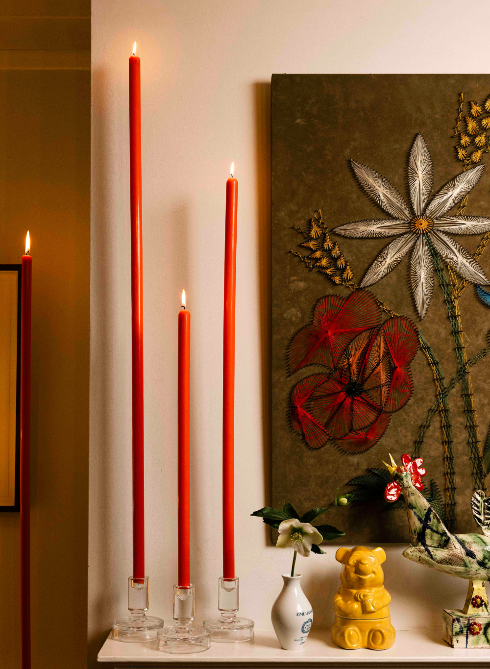 Red metre long candles in glass holders on a shelf with a floral painting in the background.