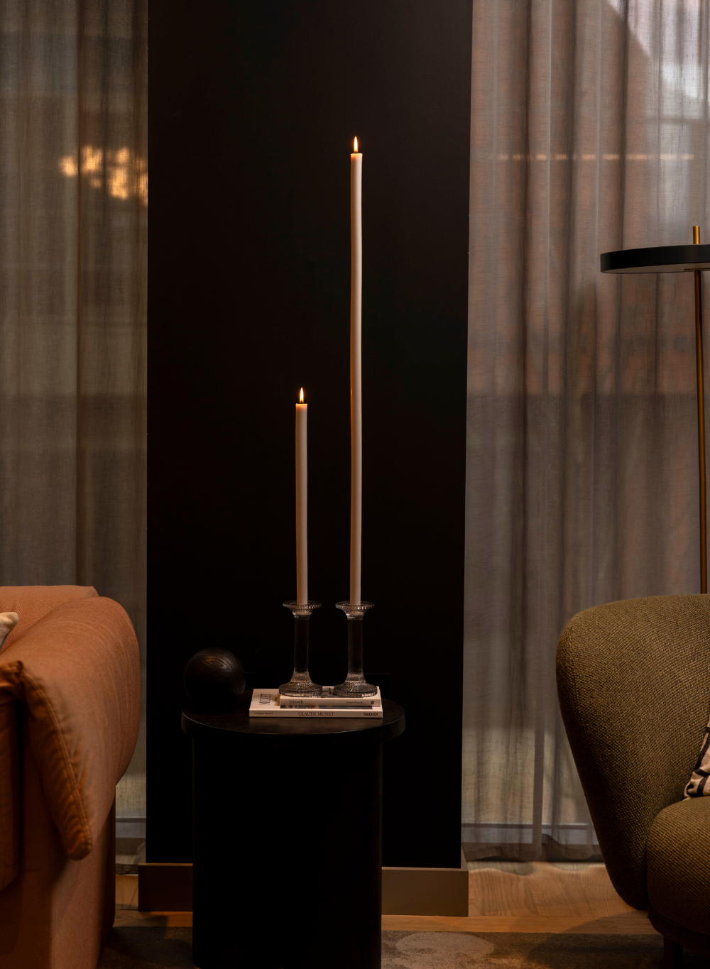 Two tall candles on a black pedestal in a dimly lit room with curtains.