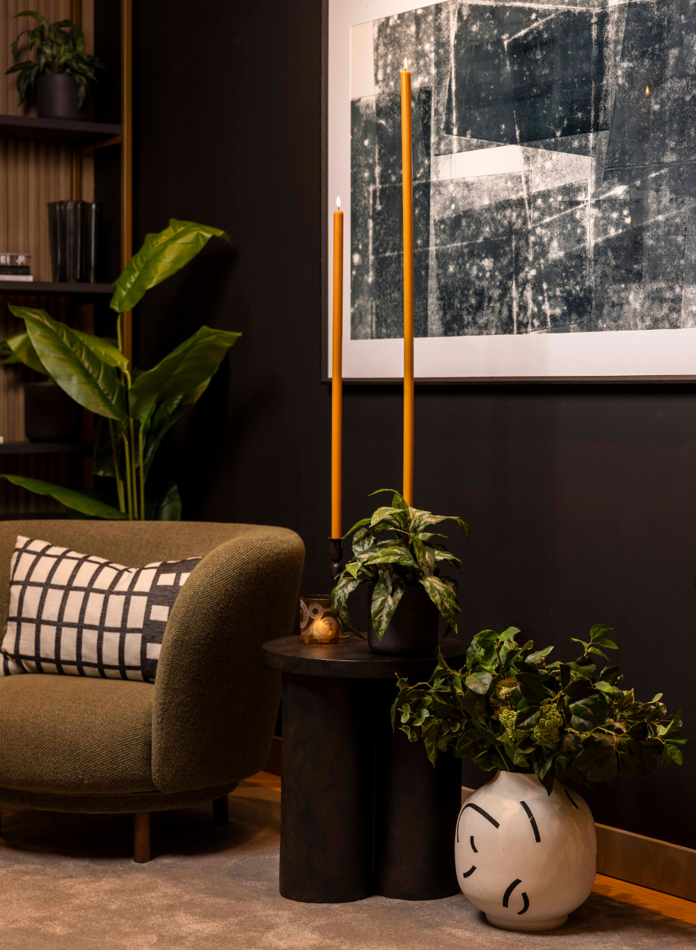 Modern interior with a green armchair, plant, and abstract art on a dark wall with metre long tumeric candles