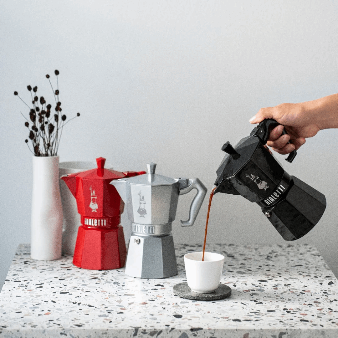 Person pouring coffee from a black Moka pot into a white cup on a speckled countertop with red and silver Moka pots.