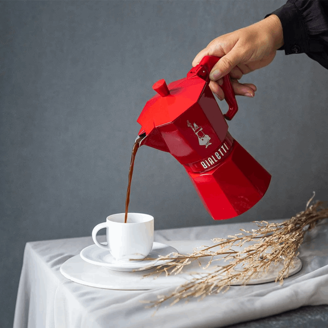 Red Bialetti coffee maker pouring coffee into a white cup on a grey background