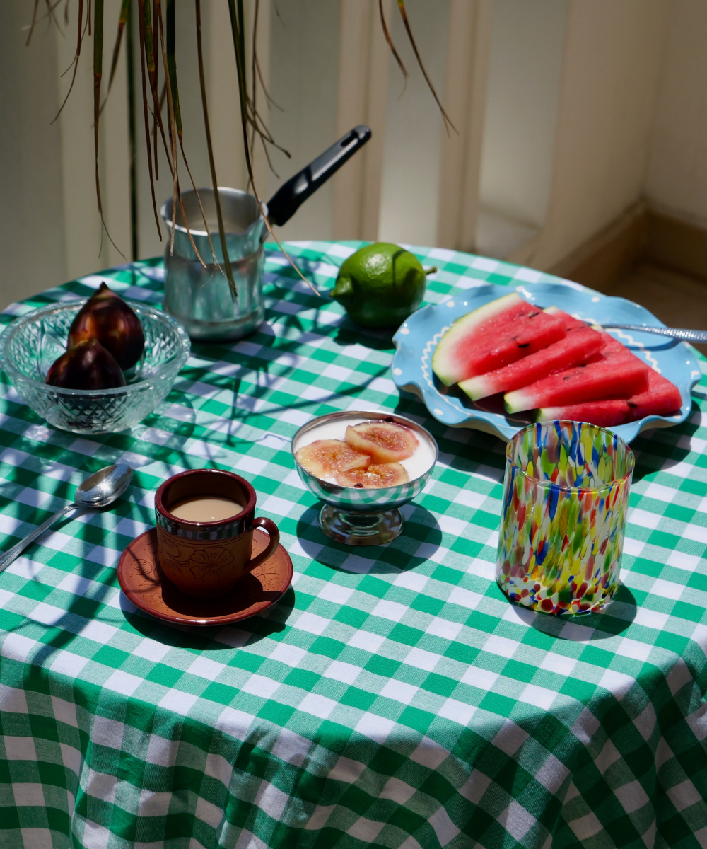 Table setting with a green and white checkered tablecloth, including a cup of coffee, watermelon slices, figs, and a colourful murano glass tumbler.