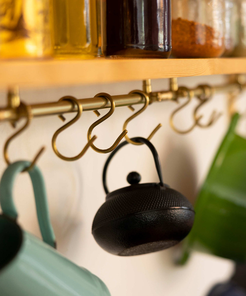 A brass hanging hook rack mounted on a wooden shelf, with a black teapot hanging from one hook in a kitchen setting. Other colorful teapots and glass jars are partially visible in the background.