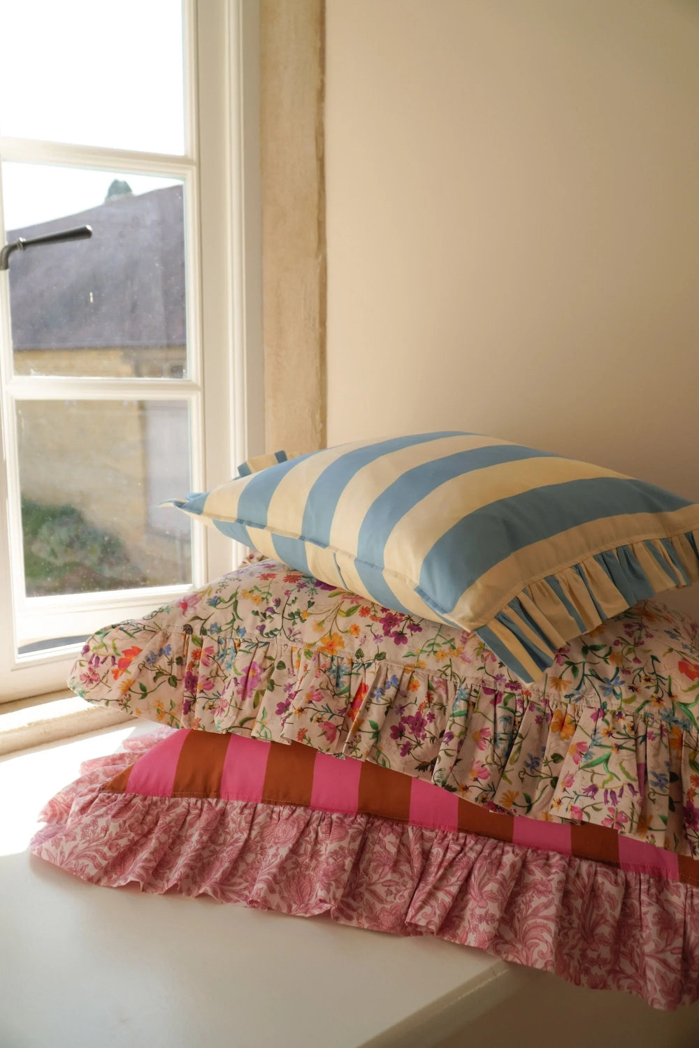 Three colourful pillows stacked on a windowsill with a view outside.