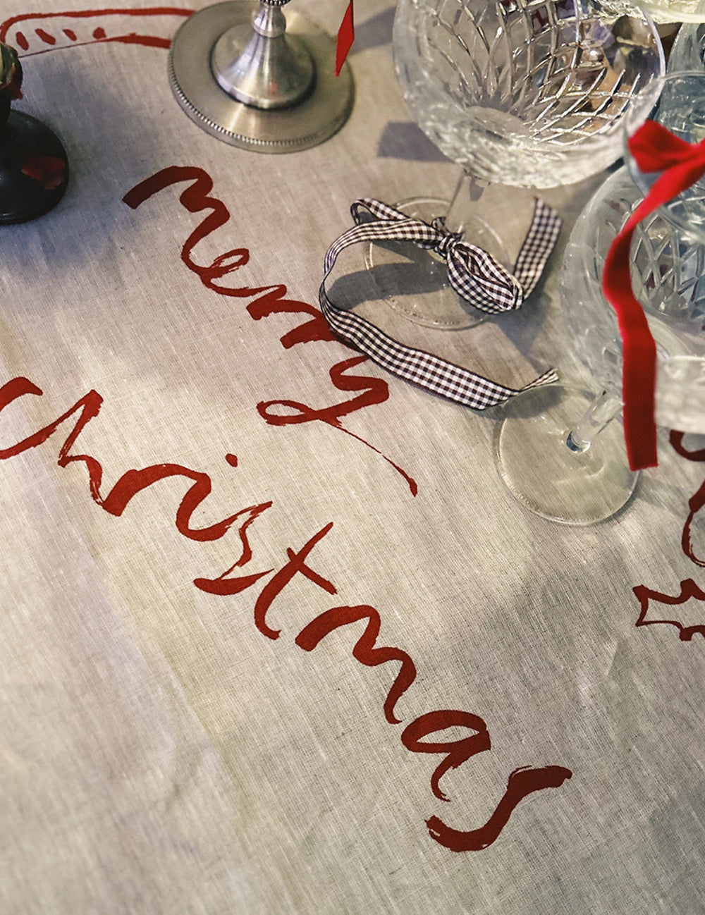 Table linen with 'Merry Christmas' text on a tablecloth, wine glasses, and decorative ribbons.