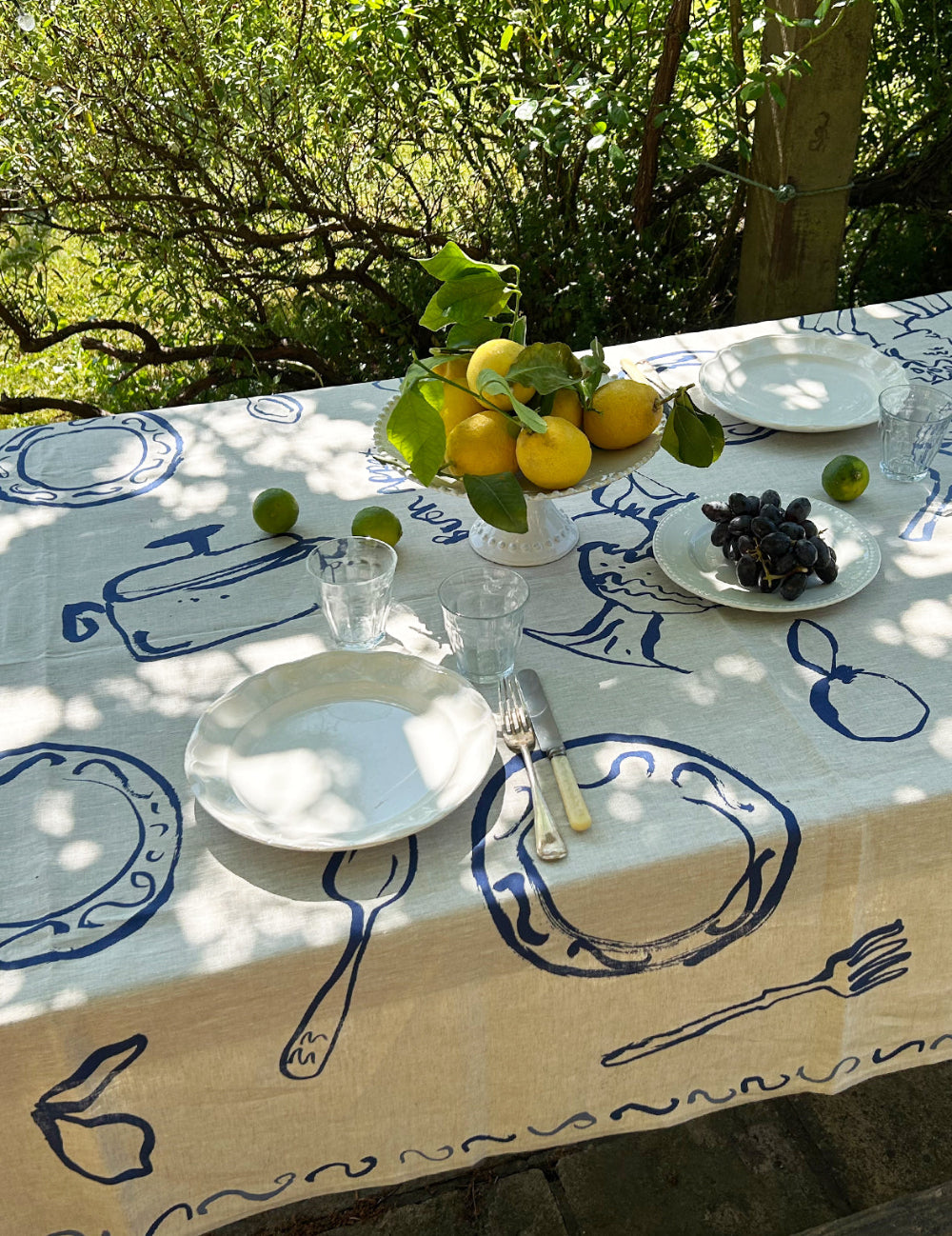 Table setting with lemons, grapes, and cutlery on a tablecloth with blue patterns outdoors.