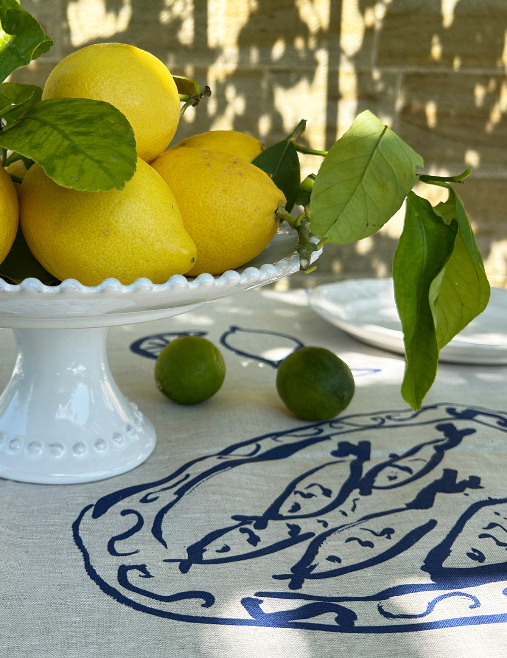 Lemons with green leaves on a white pedestal dish on a tablecloth with a fish pattern.