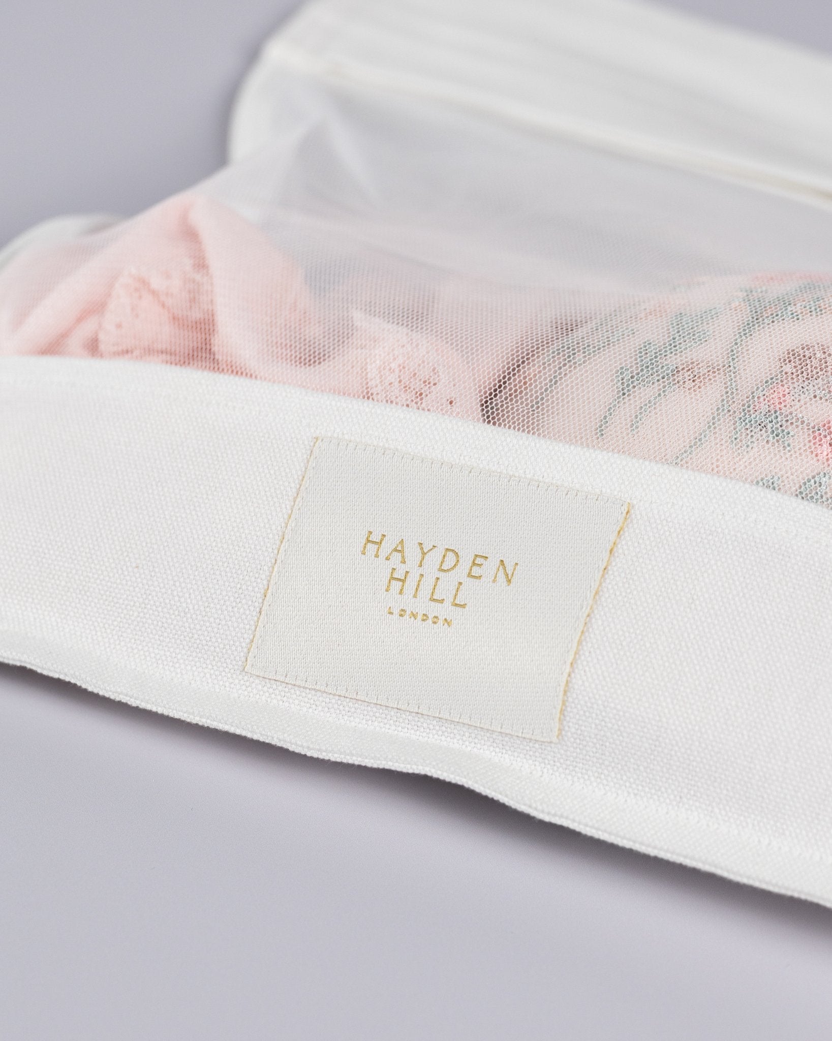 A close-up of a white Hayden Hill London garment bag with a gold-embossed logo patch, containing light pink embroidered lingerie or bedding inside.