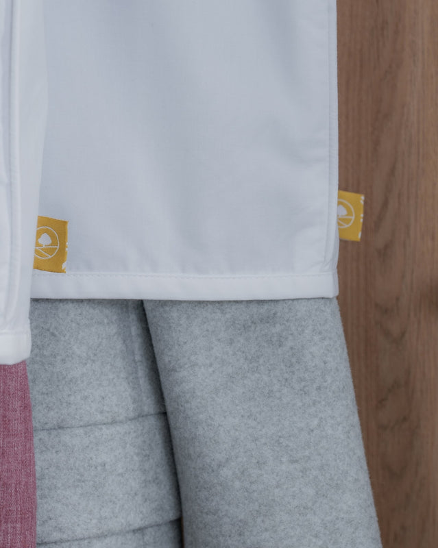 Close-up of a white garment with a hemmed edge, showing two small yellow fabric tags with a circular logo, paired with heather gray leggings or pants and a pink fabric edge on the left, against a wood panel background.
