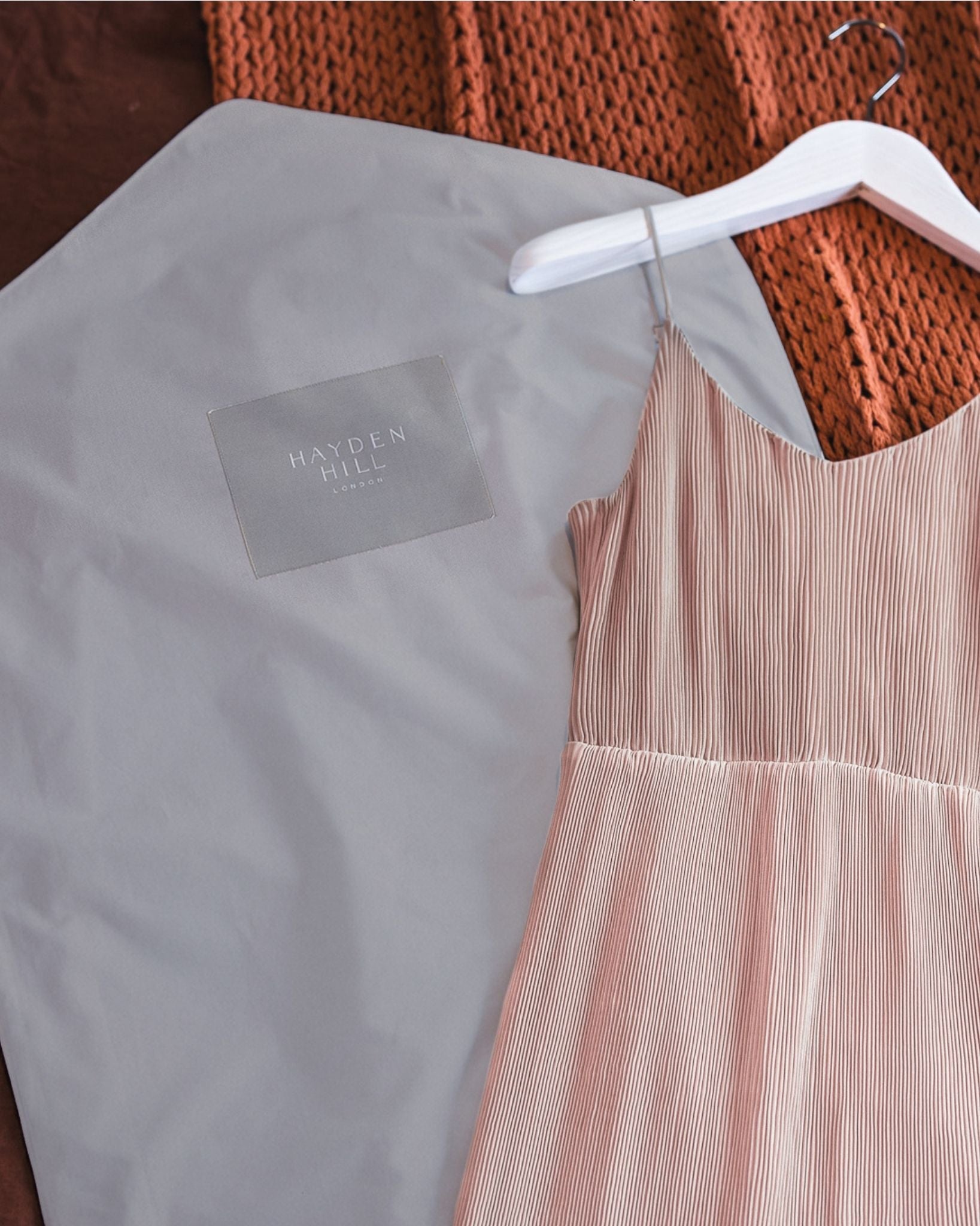 A pale blush pink pleated spaghetti-strap dress laid on a white hanger next to a light gray Hayden Mull garment bag, with a rust-orange knit blanket in the background.