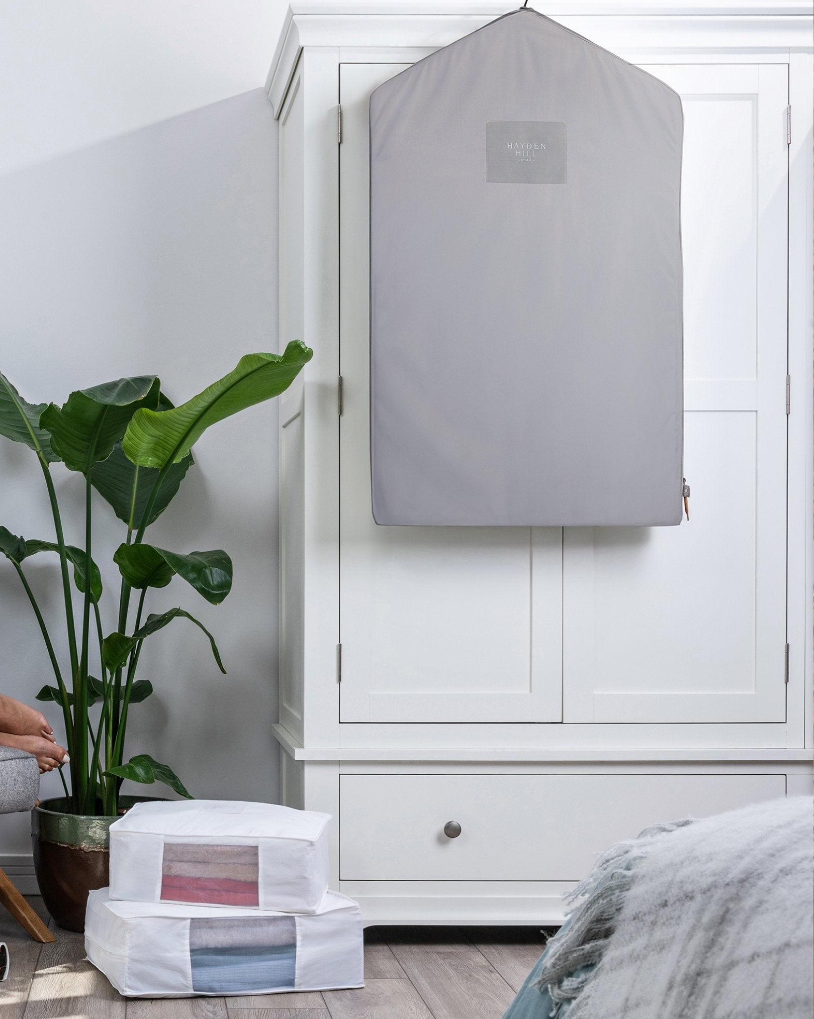 White wardrobe with a grey garment cover on a hanger, next to a plant and a bed.