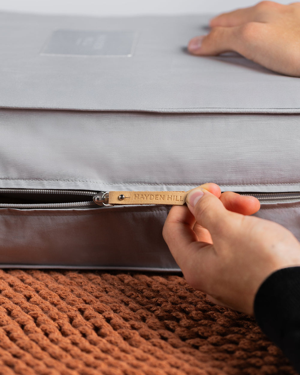 Hand zipping a grey clothes bag with 'Hayden Hill' branding on a textured brown surface.