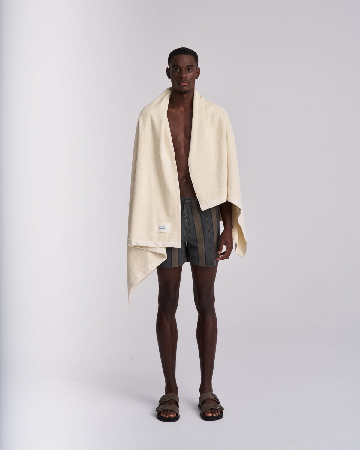 Man wearing a beige towel over his shoulders with striped swim trunks on a white background