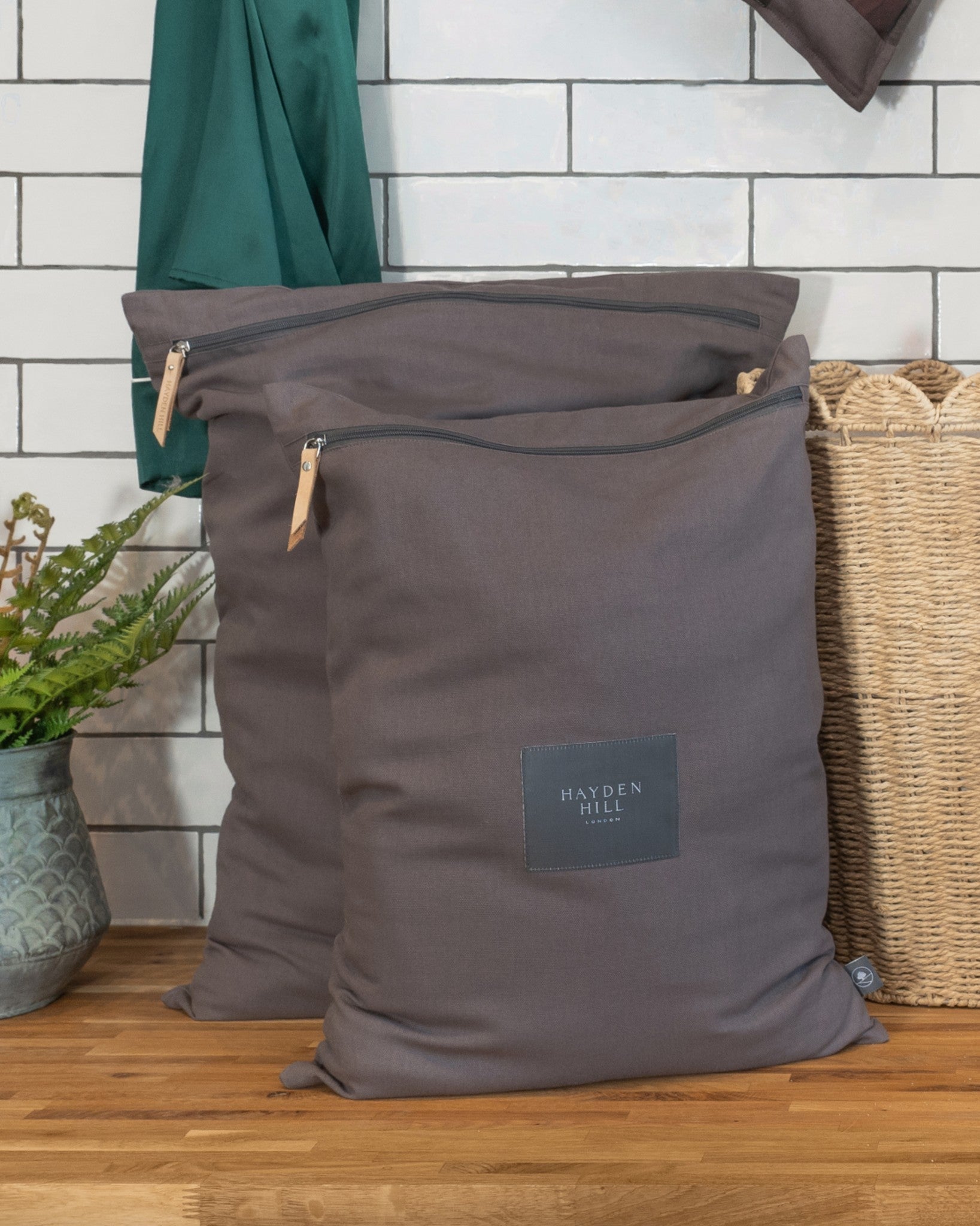 Two grey laundry bags with a visible brand logo on a wooden floor against a white tiled wall.