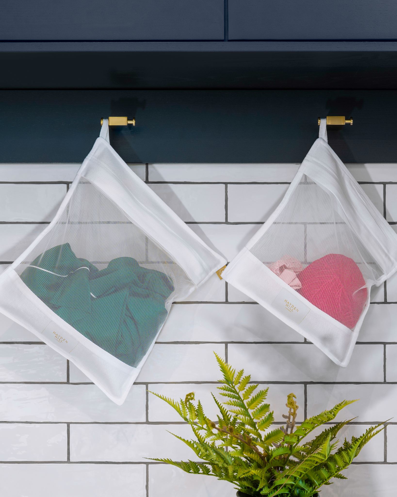 Two white mesh hanging garment bags with gold clips, one containing a green top and the other a pink knit item, displayed against a white subway tile wall with dark blue cabinets above and a green plant below.