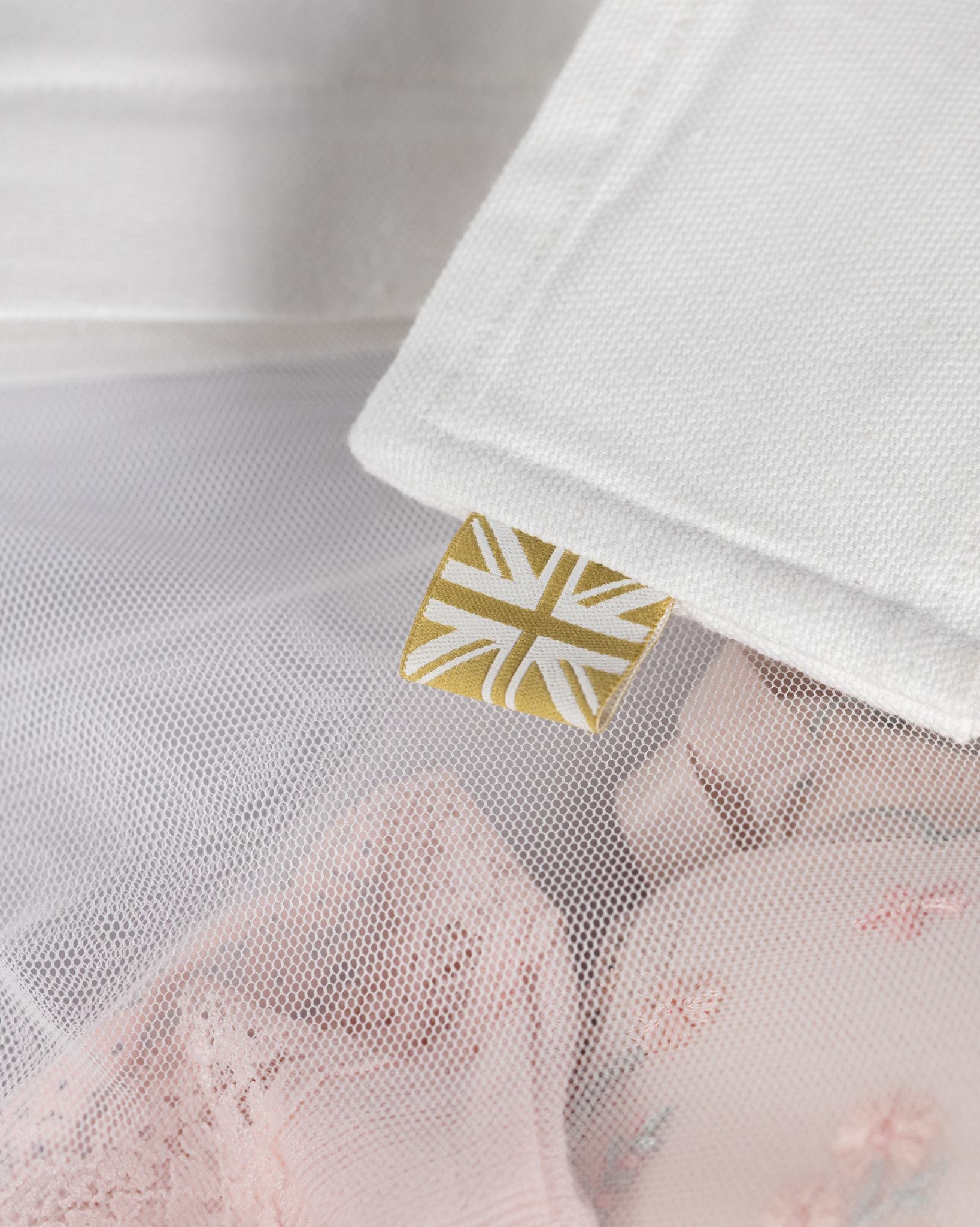Close-up of a white textured organic cotton laundry bag, with a small gold square tag featuring a Union Jack design. A soft pink lace baby garment is partially visible underneath through a sheer mesh layer.