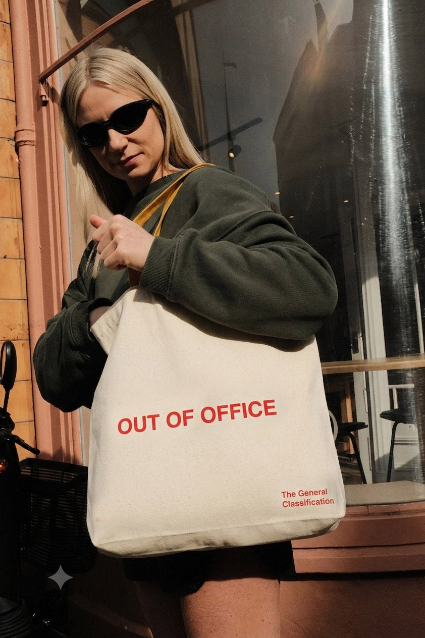 A woman wearing sunglasses and a dark green sweatshirt carries a cream canvas tote bag with red text reading 'OUT OF OFFICE' and smaller text 'The General Classification' on a city street.