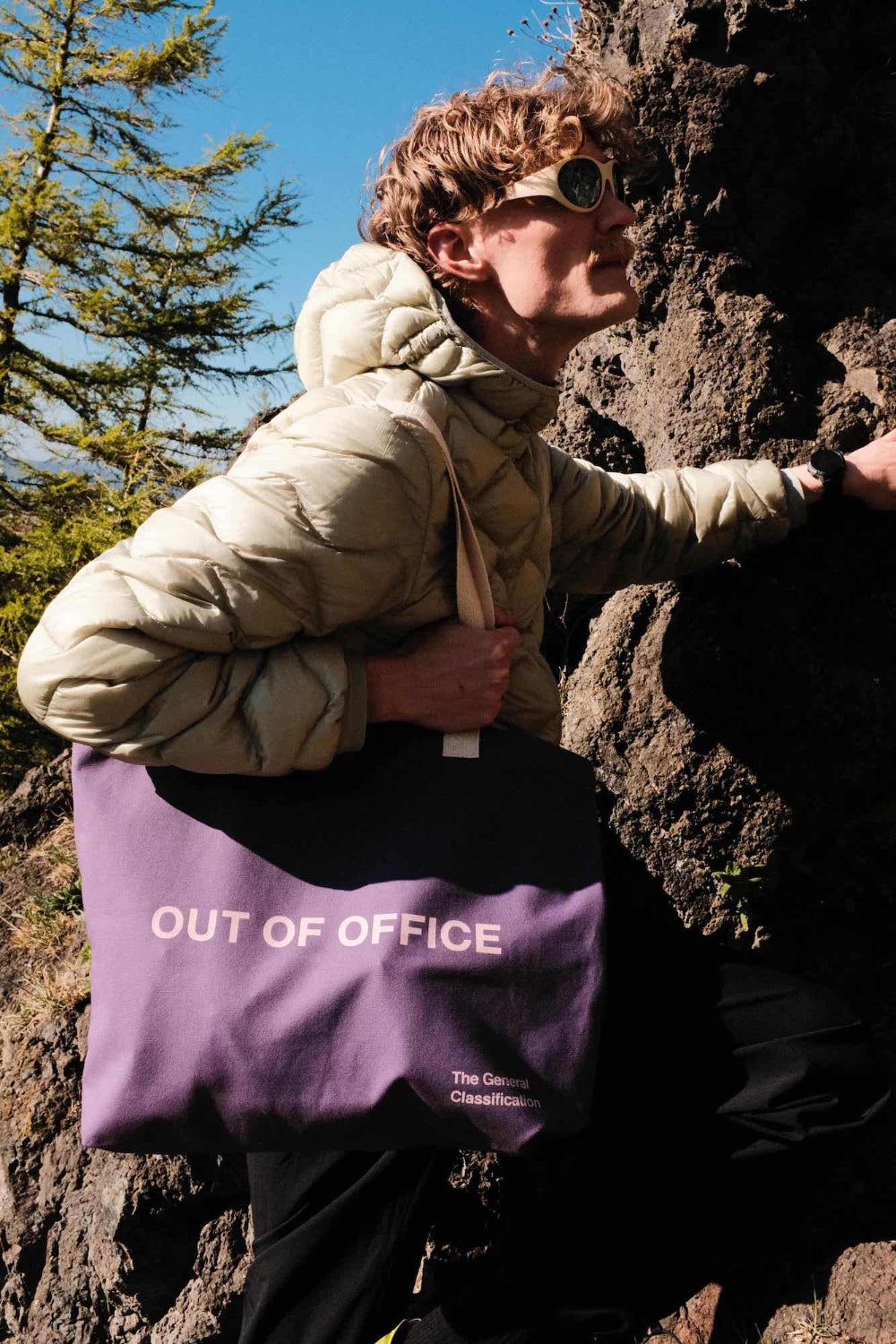 Person in beige jacket holding a purple 'OUT OF OFFICE' bag against a rocky background