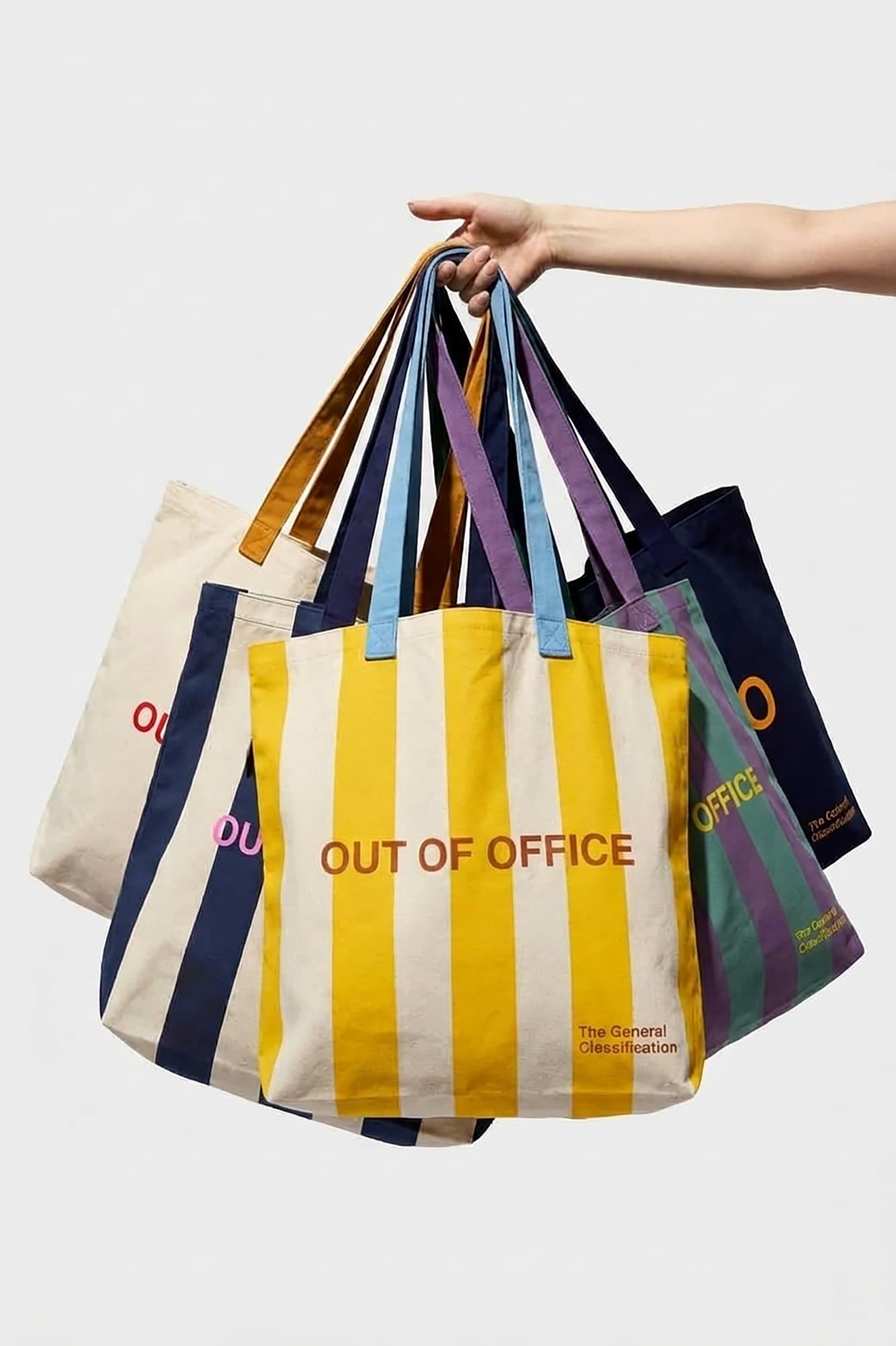 A hand holds a cluster of colorful tote bags against a light gray background. The front bag is yellow and white vertically striped with the words 'OUT OF OFFICE' in red, while the other bags behind it show navy, beige, purple, and green panels with matching branding.