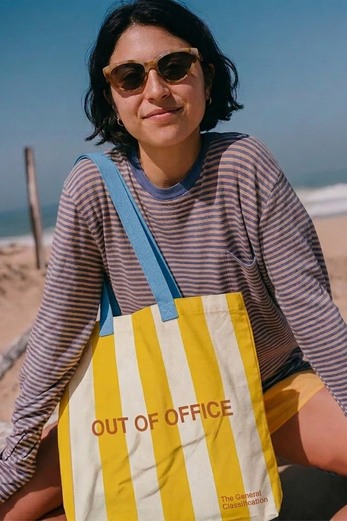 A woman wearing sunglasses sits on a beach holding a yellow and white striped tote bag with the words 'OUT OF OFFICE' printed on it. The bag has blue shoulder straps and a small 'The General classification' logo near the bottom right corner.