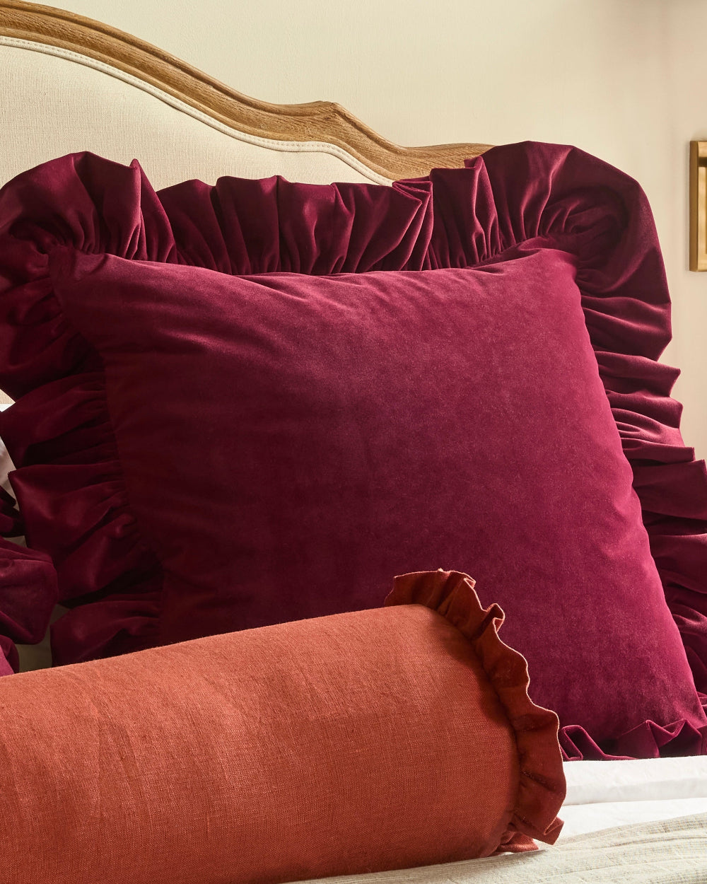 A deep burgundy velvet pillow with ruffled trim displayed on a bed, alongside a rust-orange linen pillow with a matching ruffled edge.