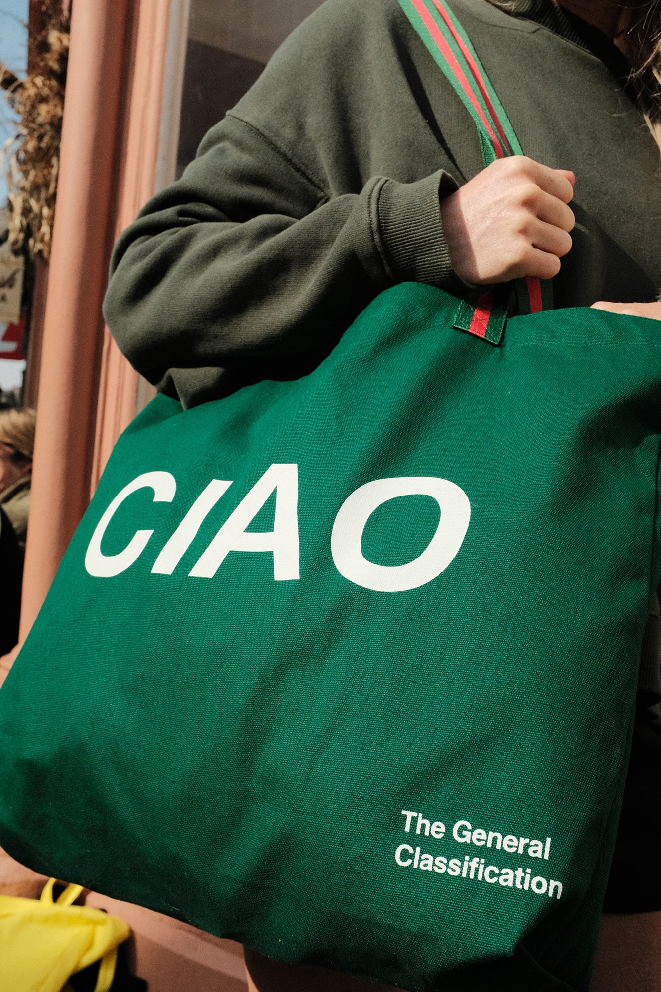 A person wearing a dark green sweatshirt carries a large green canvas tote bag with 'CIAO' printed in bold white letters and 'The General Classification' near the bottom. The bag has a green and red striped handle and is photographed in an outdoor setting.