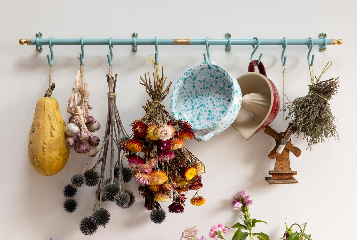 A light blue wall-mounted hanging rack with metal hooks, styled with dried flowers, garlic, a yellow squash, ceramic mugs, and a small wooden figurine against a white wall.
