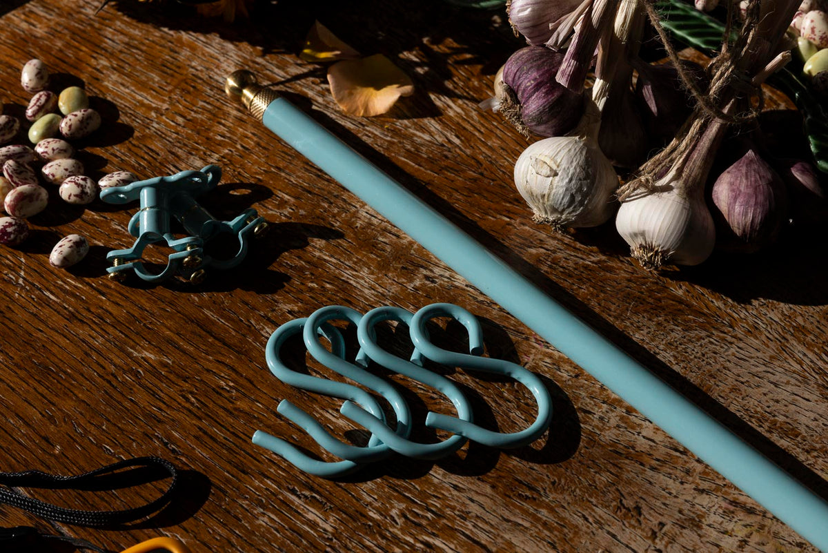 A turquoise metal curtain rod with a gold finial and matching S-hooks, displayed on a rustic wooden table with garlic and dried beans as props.