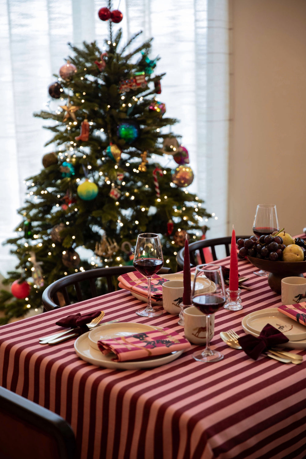 A festive dining table set with a red and white striped tablecloth, wine glasses, plates, folded napkins, and candles, with a decorated Christmas tree in the background.