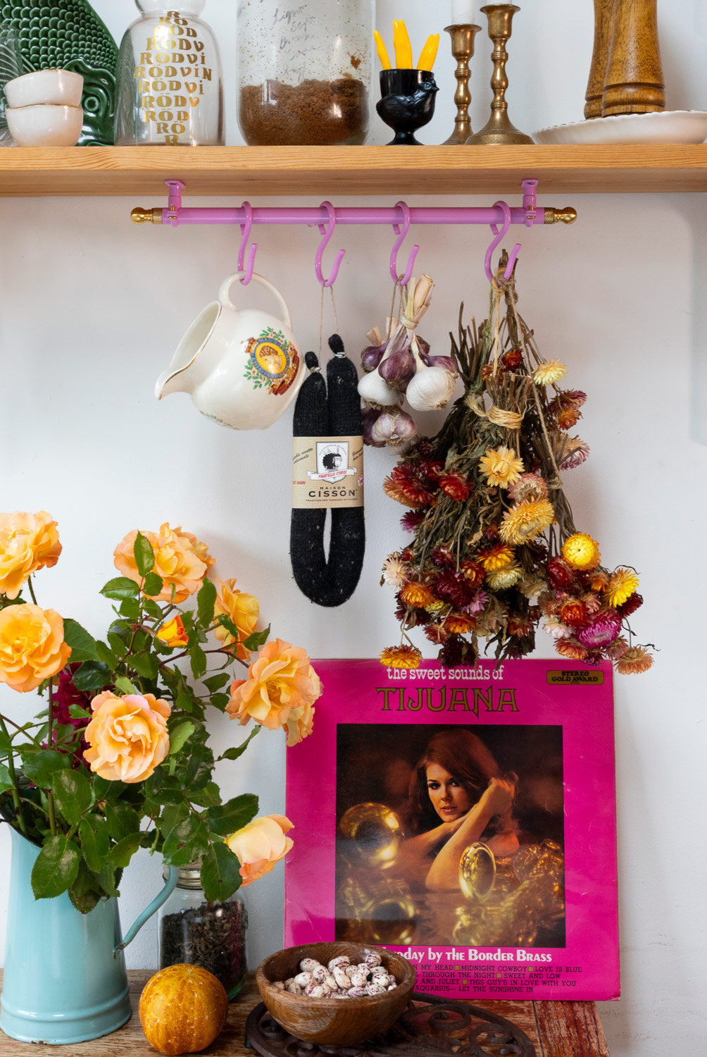 A styled interior scene with a wooden shelf and wall holding a pink hanging rack with purple hooks. A white ceramic pitcher, a black hanging item with a label, dried flowers, and a bright pink record sleeve are arranged below, alongside a blue pitcher of orange roses and other home decor.