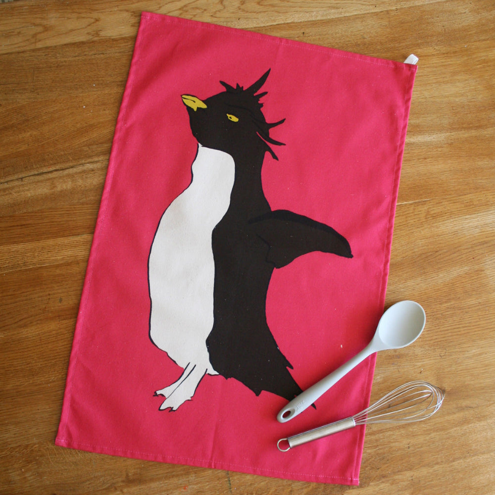 Pink tea towel with a penguin design on a wooden surface with a spoon and whisk.