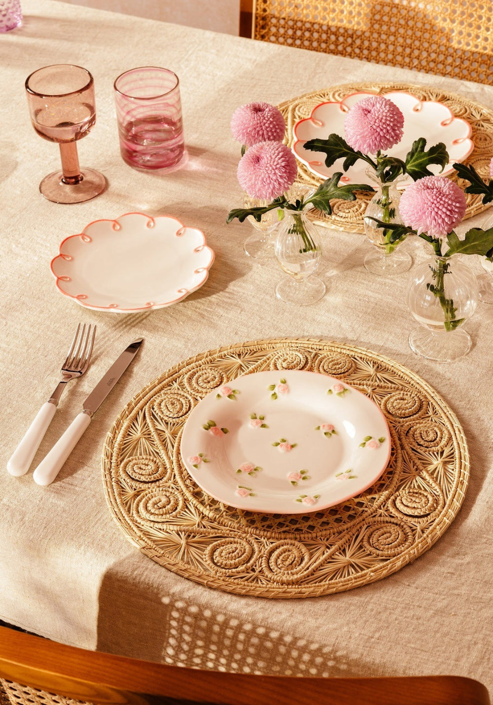 Dining table setting with decorative plates, glasses, and flowers on a light surface.