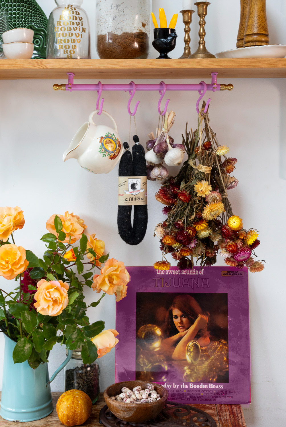 A styled interior scene with a wooden shelf and wall holding a purple hanging rack with colorful hooks. A white pitcher, a black bag, and a bundle of dried flowers hang from the rack.