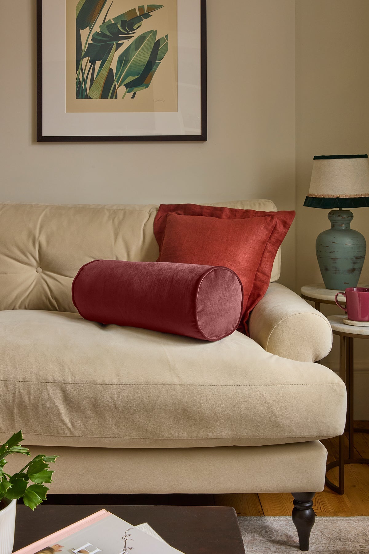 Beige sofa with red cylindrical and square pillows, framed artwork on the wall, and a side table with a lamp.
