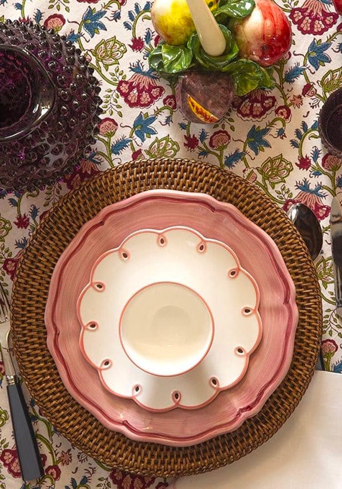 Pink ceramic plate with scalloped edge on a woven placemat over a floral tablecloth.