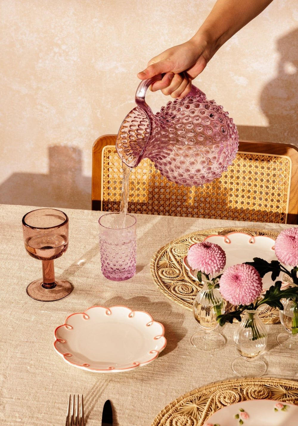 Person pouring water from a decorative glass jug into a glass on a table setting with flowers and plates.