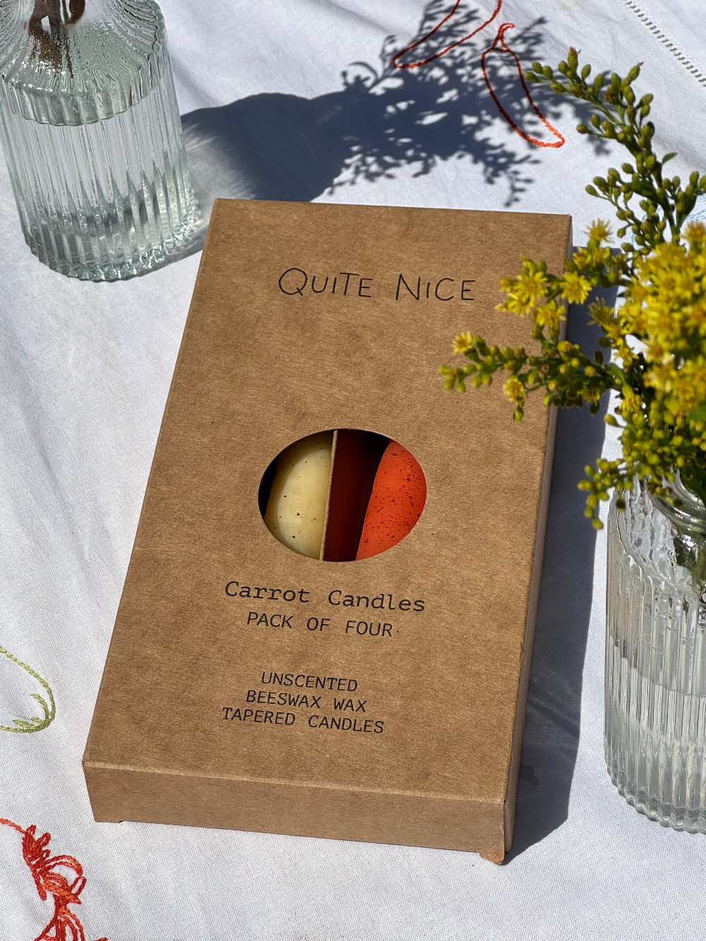 Quite Nice London rainbow carrot candle gift box displayed on white embroidered table linen beside yellow flowers and glass vases.