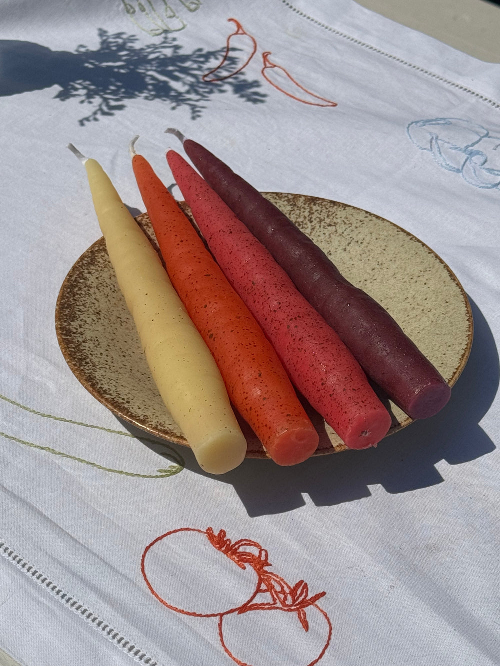 Four unlit carrot-shaped beeswax candles in yellow, orange, pink and burgundy arranged on a speckled ceramic plate.
