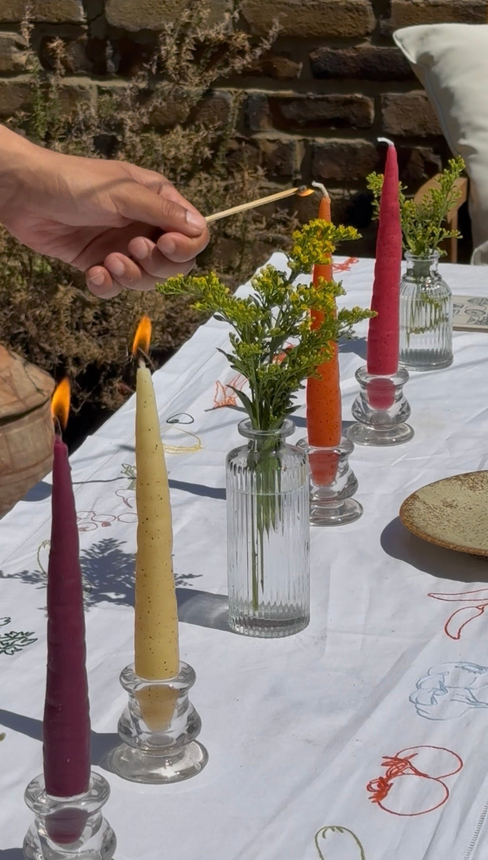 Colourful carrot candles displayed in glass holders on an outdoor dining table with flowers, as one candle is being lit.
